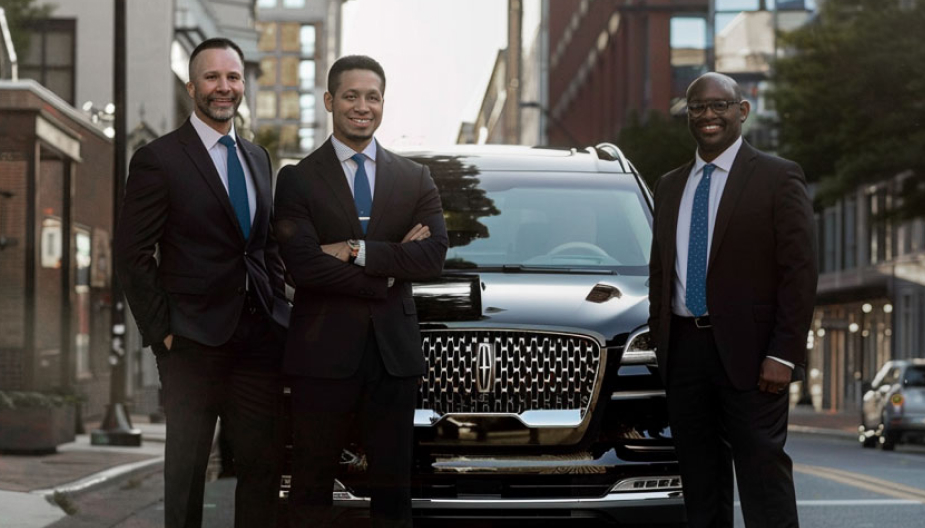 Men in black suits and blue ties in front of a luxury vehicle.